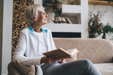 Cheerful senior woman sitting alone and reading book in free time at home