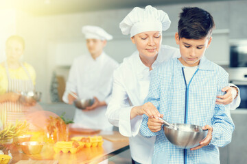 In restaurant kitchen during children cooking class, female chef help teen boy thoroughly stir homogeneous liquid dough with whisk. In background, blurry participants are standing near table with food