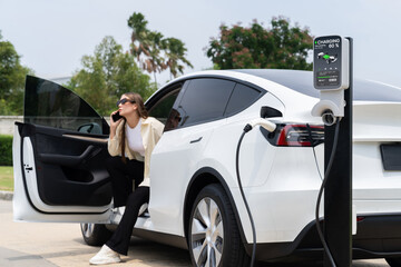 Young woman recharge her EV electric vehicle at green city park parking lot while talking on phone....