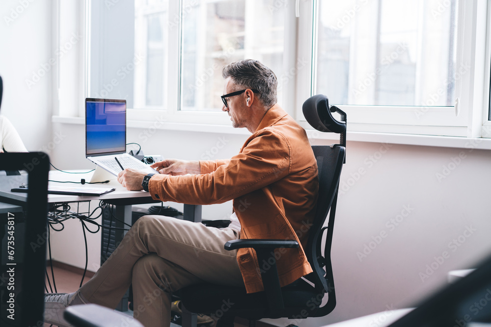 Poster side view of skilled entrepreneur in bluetooth earbuds browsing web page on modern laptop computer d