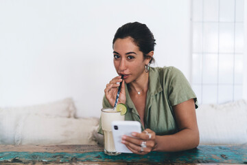 Positive ethnic woman sitting at table and drinking glass of milkshake at cafe