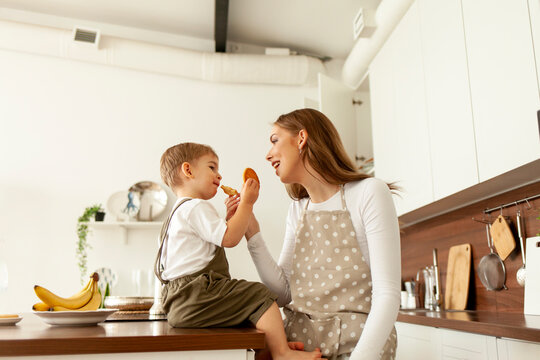 mother in apron feeds child in the kitchen, young woman cook gives cookies to her little son to eat at home