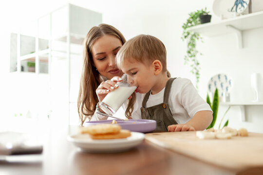 mother feeds her little son at home in the kitchen, woman gives glass of milk for breakfast, hungry boy drinks milk