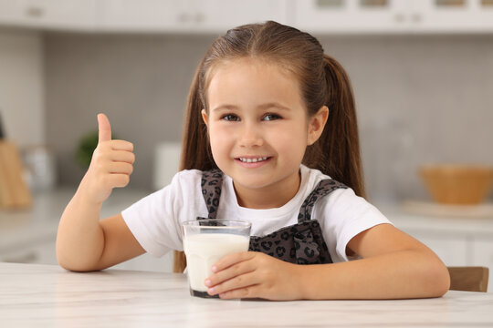 Cute girl with glass of fresh milk showing thumb up at white table in kitchen