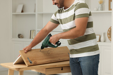 Man with electric screwdriver assembling furniture at table in room, closeup