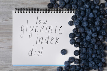 Notebook with words Low Glycemic Index Diet and blueberries on light wooden table, top view