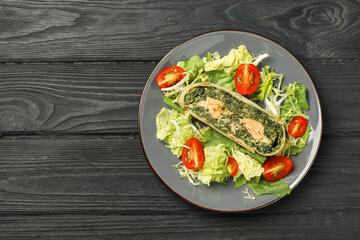 Piece of tasty strudel with salmon, spinach and salad on black wooden table, top view. Space for text