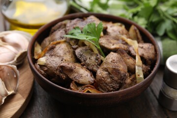 Tasty fried chicken liver with onion and parsley in bowl on wooden table, closeup