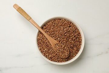 Bowl and spoon with dry buckwheat on white table, top view