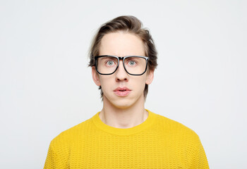 A young man in a yellow sweater and glasses poses on a gray background.