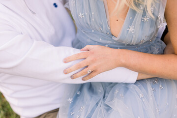 An engaged couple in low sits together, he has his arm around her waist and she shows off her engagement ring while touch his arm. She wears a blue dress, and he wears a white shirt.