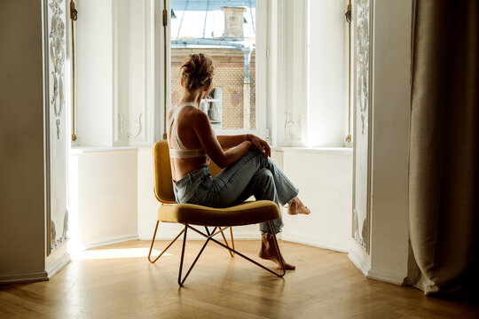 A beautiful blonde woman in jeans and a bra sits in an armchair looking out the window