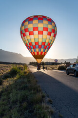 A colorful hot air balloon lands on a road in Albuquerque, New Mexico during the Balloon Fiesta. The balloon is lit like stained glass by the morning sun. The road leads toward the mountains behind.