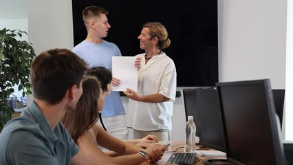 Positive male tutor presenting certificate confirming successful completion of computer courses to satisfied young guy in front of student group in classroom