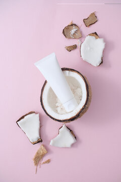 A lotion tube without label is placed inside half a coconut on a pink background. View from above. Coconut water contains cytokinin, which helps fight skin aging. Mockup, flat lay.