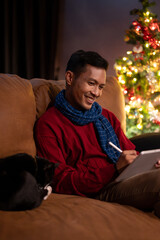 A happy Asian man is using his digital tablet on a couch while his lovely cat is sleeping beside him