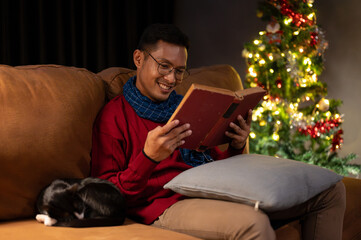 A happy Asian man is reading a book on a couch while his cat is sleeping beside him.