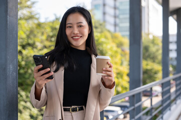 An attractive Asian businesswoman is holding a coffee cup and her phone while standing on a skywalk.