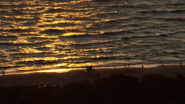 Palanga beach by the Baltic sea. Beautifully lit by sunset light. 7x zoom aerial shot. Waves and the people silhouette on the beach are visible.