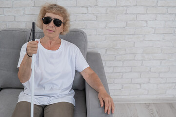 An elderly blind woman wearing sunglasses and with a tactile cane sits on the sofa. 