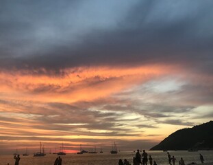 sunset over the mountains with so many yachts above the sea and the silhouette of tourists on the beach