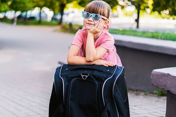 Close-up of a cute happy little traveler girl in sunglasses leaning on a black suitcase. Smiling pretty small girl with a suitcase. Children go abroad for summer holidays or weekends. Travel concept