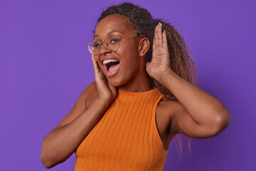 Young cheerful excited African American woman is surprised and puts palm to ear to hear answer to question or new shocking news dressed in casual clothes stands on plain purple background.