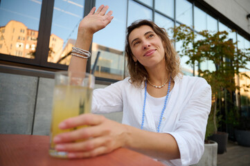 Young woman in a white shirt and a necklace around her neck on a bright sunny day in a cafe with greenery in the background is drinking fresh juice and actively talking about something