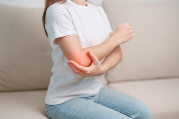 Woman having elbow ache during sitting on couch at home, muscle pain due to lateral epicondylitis or tennis elbow. injury, Health and medical concept - obrazy, fototapety, plakaty