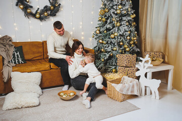 A cheerful young family with a small child rests near the Christmas tree at home. Family couple with Christmas decoration at home. Christmas, New Year - time to celebrate