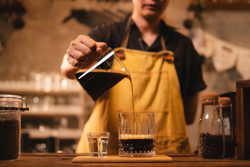 A female barista is pouring an americano or filter coffee to a glass. Concept of vintage cafe or coffee shop. Close up of hand pour black coffee ready to serve. Preparation, aroma and smoke.