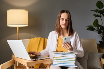 Concentrated focused woman using smart phone and her notes for preparing for test doing homework...