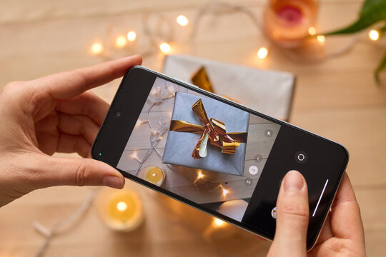Unrecognizable woman making photo with her smartphone photographing present box with lights and candles decoration creating festive content for social network.