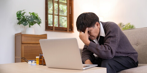 Asian Mature adult man suffering from fever, calling his doctor and touching his head, sitting on couch in front of laptop in living room