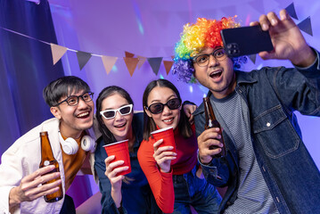 Fototapeta premium Happy asian diverse group friends taking selfie with neon light on a party using smartphone. Home party friends having fun enjoying time and dancing together. New Year's Eve Party concept