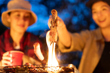 Close up of asian woman friends enjoy drinking a beer and grilling meat together. They feeling exited while camping in the nature at night. Recreation and journey outdoor activity lifestyle.