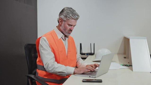 Serious caucasian man male engineer architect with grey hair dressed in high visibility orange vest sitting at workplace and typing on wireless laptop. Concentrated worker creating business project.