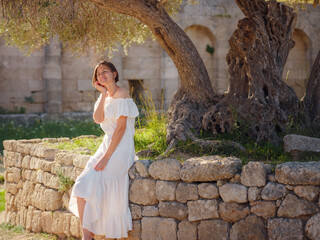 Beautiful Asian young woman in white dress outdoor. Acropolis of Rhodes Famous ruins of ancient settlement with various buildings such as stadium and theater.