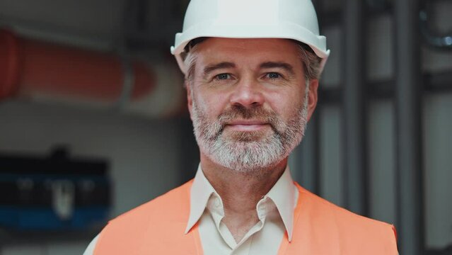 Portrait of handsome mature male engineer posing on background of metal pipes with flowing water. Bearded man plumber with hard hat showing competence with serious expression on face. Boiler room.