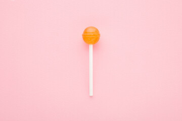 Orange lollipop on stick on light pink table background. Pastel color. Sweet candy. Closeup. Top down view.