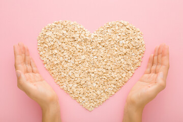 Young adult woman hands and heart shape created from dry rolled oat on light pink table background....