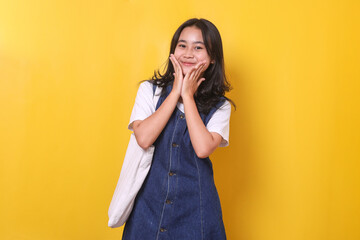 Cute young Asian student girl holding cheeks looking at camera on yellow background