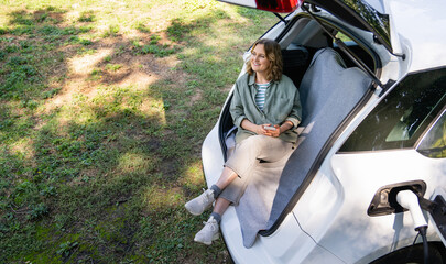 Woman with smartphone sits in an electric car's trunk