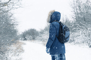 Back view of traveler in a blue jacket with a fur hood and a backpack on the background of a winter landscape during snowfall.