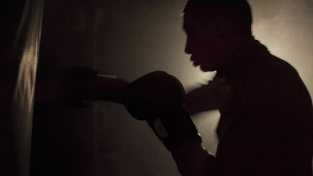 Male boxer training with punching bag in dark sports hall. Exercise for the big fight. Boxer hits