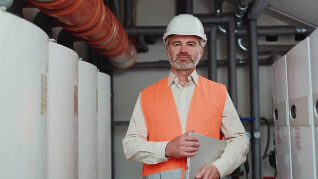 Proud male engineer in orange safety vest standing with closed laptop in hands on workshop background. Handsome elderly male looking at camera after providing quality control at modern factory.