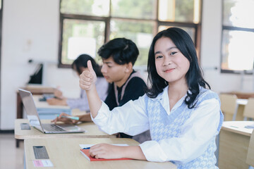 Portrait of smiling asian female student sitting while giving thumb up  at desk in classroom at university