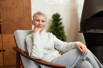 Elegant confident senior female in stylish gray sweater sitting in chair leaning elbow on armrest putting hand under chin, looking at camera, relaxing next to chimney in cold winter day - obrazy, fototapety, plakaty