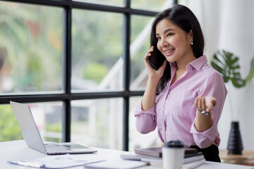 Asian Woman entrepreneur busy with her work in the office. Young Asian woman talking over smartphone or cellphone while working on computer at her desk.
