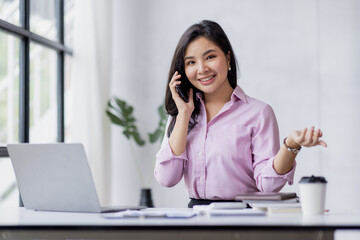 Asian Woman entrepreneur busy with her work in the office. Young Asian woman talking over smartphone or cellphone while working on computer at her desk.
